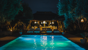 Looking across a pool at night to the nice outdoor kitchen and dining area on the other side.