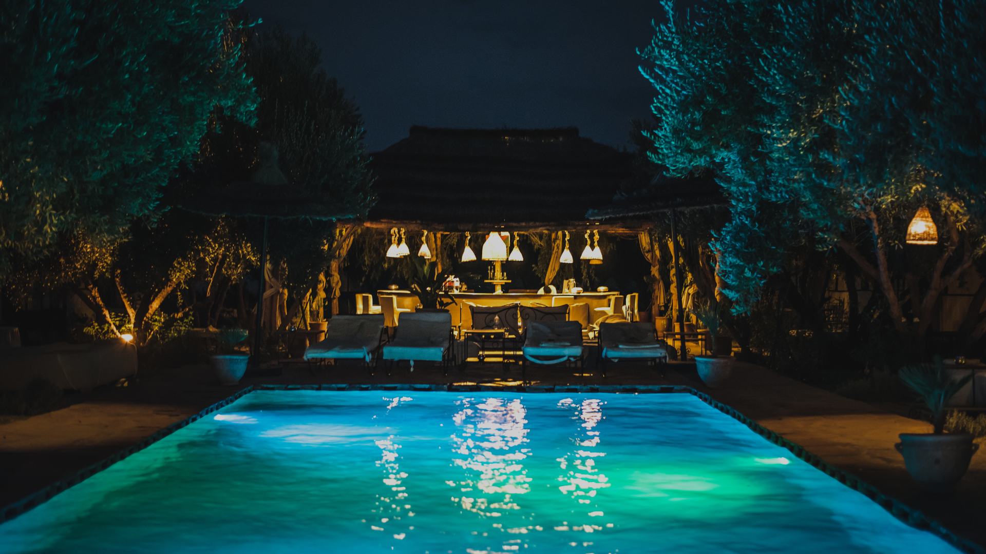 Looking across a pool at night to the nice outdoor kitchen and dining area on the other side.